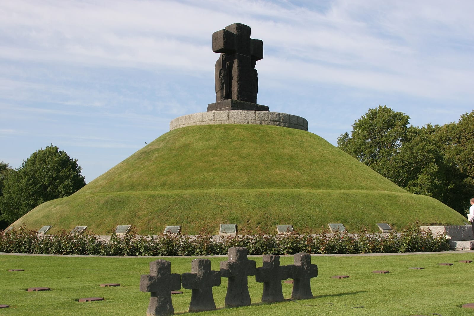 Tumulus herbeux surmonté d'une croix et de statues sombres, avec de petites croix de pierre au premier plan