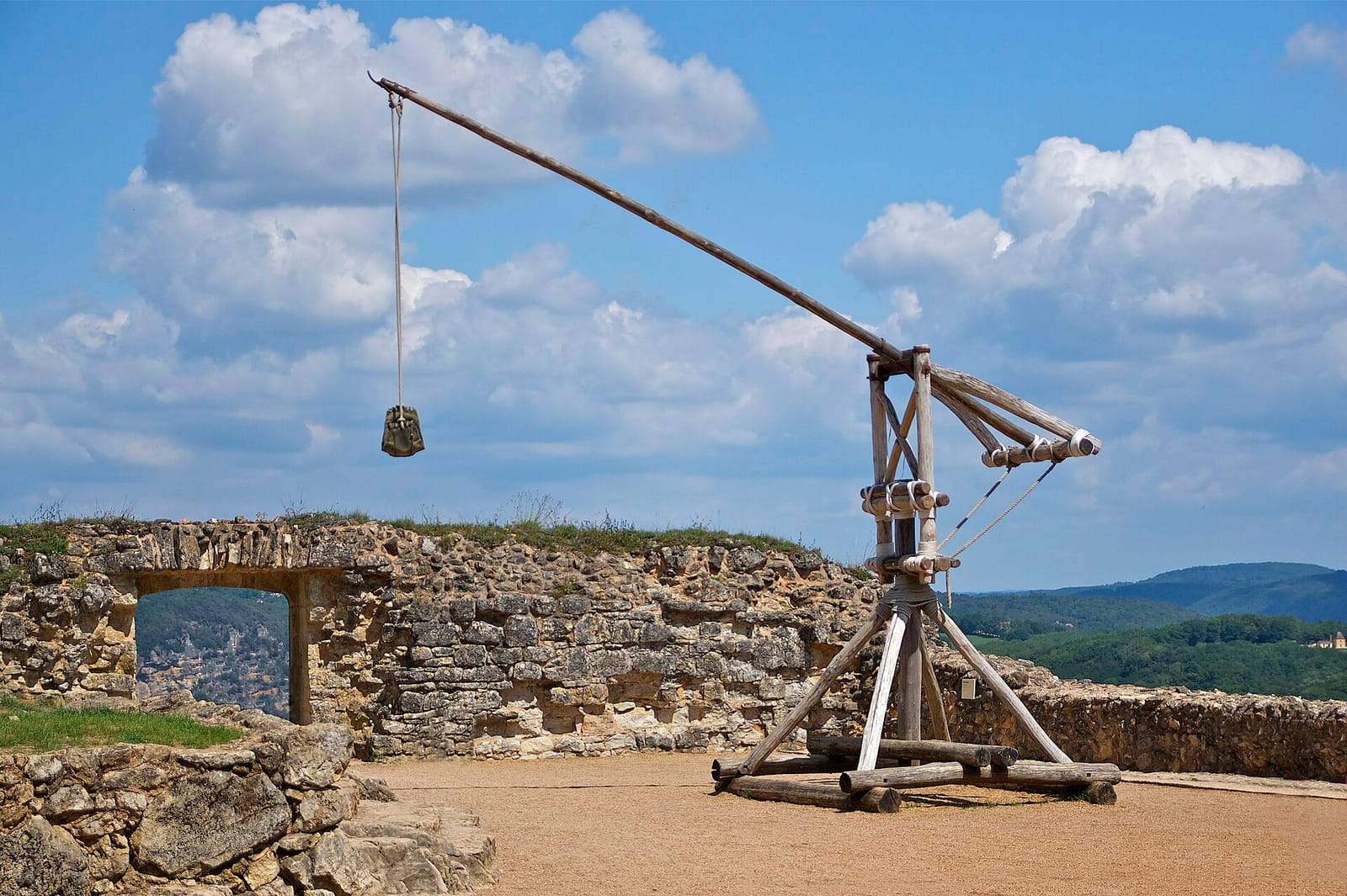 Grande machine de bois à long bras et contrepoids, dressée devant un paysage de collines